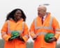 Miatta Fahnbulleh and John Healey, wearing orange hi-vis jackets and holding green safety helmets, smile as they talk and walk side by side
