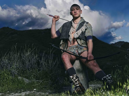 Olly Alexander sitting on a tree stump with mountains in the background, and carrying a bow and arrow