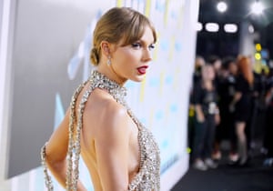 Taylor Swift poses in front of cameras. She is wearing a silver jewelled dress and is looking back over one shoulder