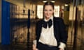 Drew Barrymore, hair parted in the middle and pulled back and wearing a satin shirt with a big bow, bracelets and a suit jacket, smiles as she stands with her hands in her trouser pockets in a school hallway with lockers