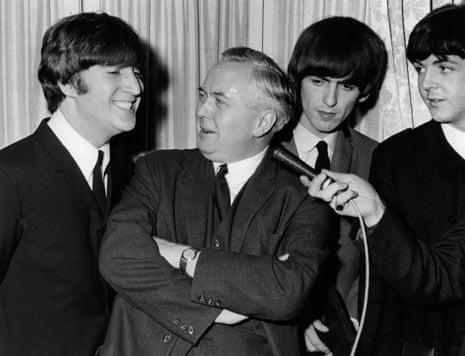 A black and white photograph of Paul McCartney holding a microphone up to Harold Wilson, who is looking at a grinning John Lennon, while George Harrison stands behind them. They are all wearing suits and ties