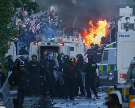 Riot police and their vehicles in the foreground, and in the background behind a large fire, a crowd