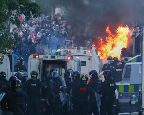 A group of police in riot gear assemble behind vans with a crowd of rioters in the background