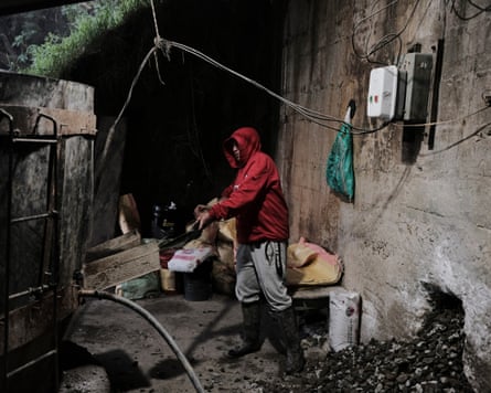 A man works in a yard with exposed wires and pipes crossing it. He is lifting a large shovel to a large wooden vat.