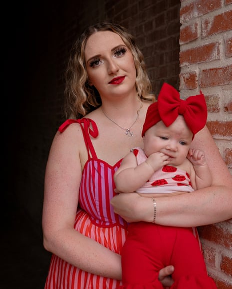 Aubrie Morris standing outiside near a brick wall, wearing a red and pink dress with tie straps, holding her baby who is dressed in a red outfit with strawberries on the top half and a red bow around her head