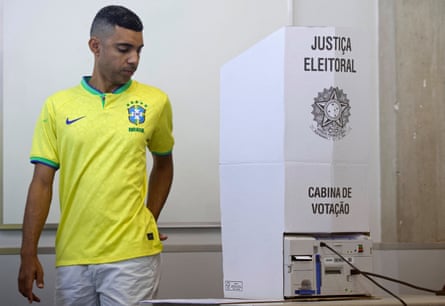 A voter in a Brazil football jersey stands beside a voting booth at a polling station
