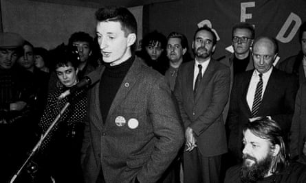 A black and white image of Billy Bragg standing at a microphone with Neil Kinnock watching him in the background