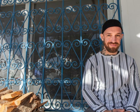 A man in a skullcap and robe sitting in front of a blue iron and a pile of wood.