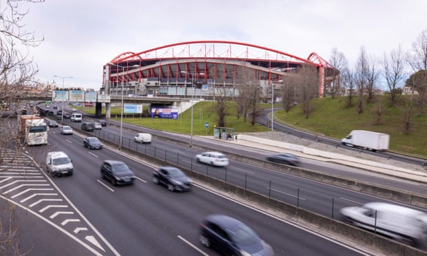 A stadium wih red roof supports, next to a motorway