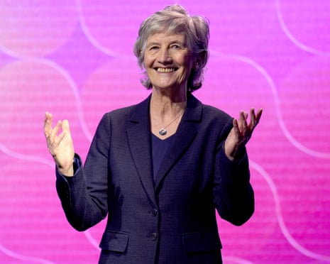 Head and shoulders picture of Catherine Connolly, wearing a dark suit and smiling, with her hands raised, against a pink background