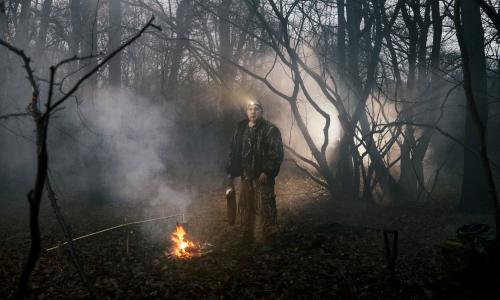 Steve Hart standing in woodland next to a campfire, holding a dead rabbit by its feet