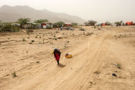 A child pulls a water can along a dirt track with tents in the background.