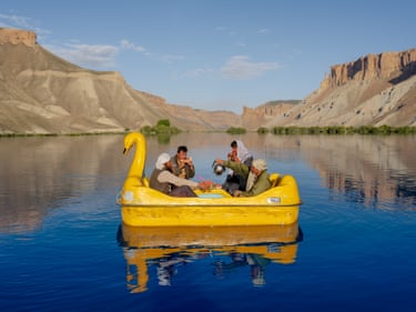 Four men drink tea and eat watermelon while on board a large yellow, swan-shaped dinghy, on a wide empty lake under a blue sky