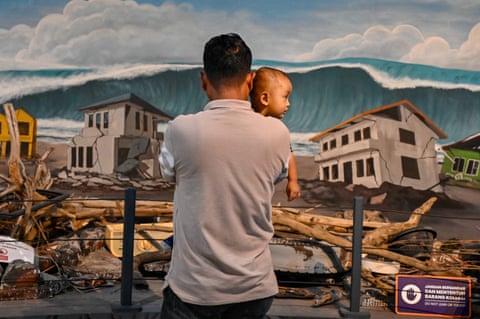 A man looking away from the camera, holding his baby, as they look at a display showing rubble and pictures of houses with a tsunami rising above them