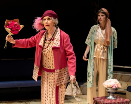 Patricia Hodge wears a matching pink patterned dress and jacket with a pink hat and is holding pink flowers. Zoe Brough stands behind her, wearing a silk scarf tied around her head and a loose light green jacket