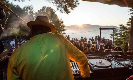 A DJ performs in front of a crowd at the Meadows in the Mountains festival, Bulgaria, in the distance the sun sets on the mountains.