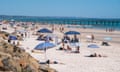 Henley beach in Adelaide on 07 Jan 2023. People are lying down and standing under umbrellas on the sand.  In the distance, there is a bridge and there are people near the water.