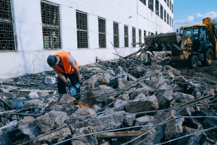 A man in a hi vis jacket saws through chunks of metal wires stuck through concrete in a pile of rubble next to a building with a digger behind him
