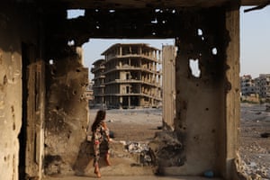 A woman sits on the window ledge of a ruined building, looking out over other ruined buildings in the distance