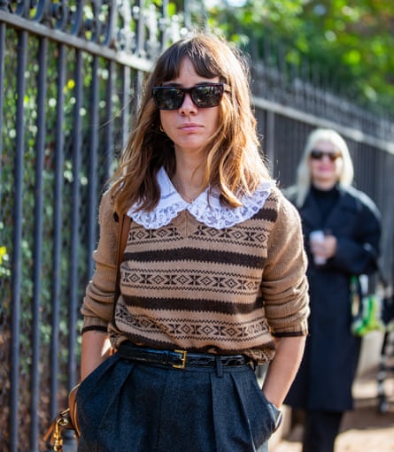 A woman with long brown hair and dark glasses, wearing a brown patterned jumper over a white blouse with a lace collar, with baggy tweed trousers, walking along a street next to railings and trees, with a blond woman behind her.