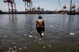 A boy jumping into water that has rubbish floating in it, with an industrial site in the background