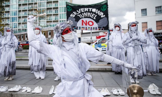 Protesters in fancy dress stand in front of an anti-tunnel banner, with pairs of white shoes laid out neatly in a row on the pavement