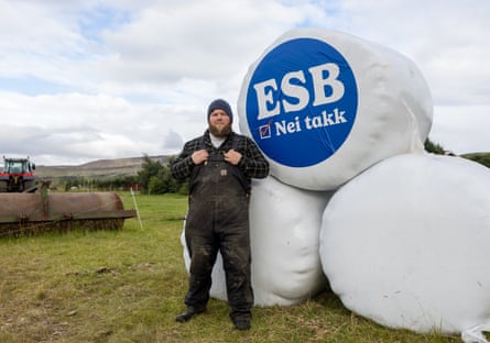 Davíð Clausen Pétursson stands beside a hay bale covered in white plastic and with a blue sign reading “ESB nei takk”. Davíð is sturdily built with a bushy light brown beard, and wears a woolly hat, checked shirt, and dark blue heavy-duty dungarees.
