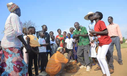 A man in a green flowered shirt sings in semicircle of onlookers while a man crouches to play a drum.
