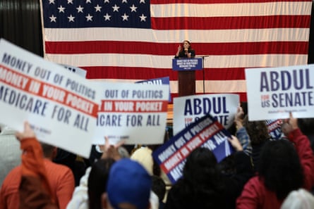 Seen from a distance, a woman stands at a lectern in front of an enormous American flag, with crowd members holding up political signs.