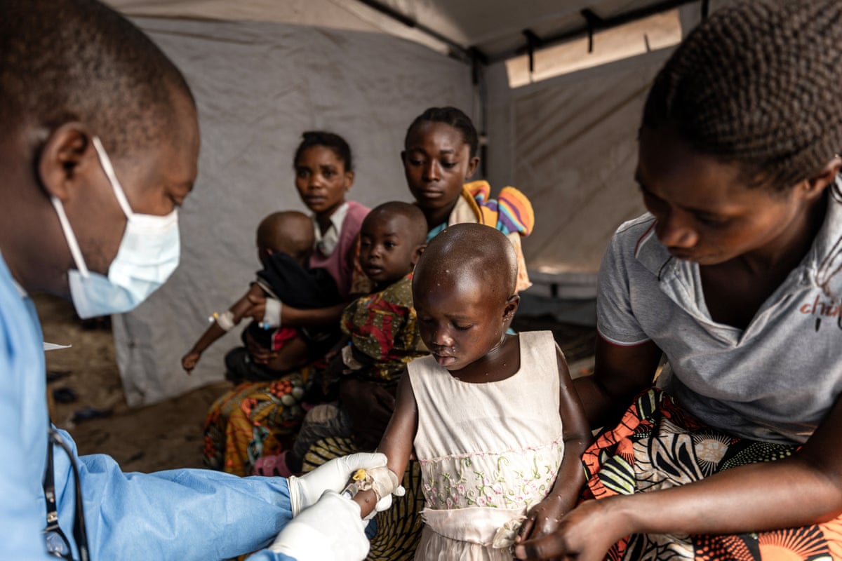 A young girl watches as a masked medic uses a canula in her arm, while two other mothers waiting with their children in the background