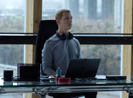 A man in a blue top, with headphones around his neck, sits very upright at a glass desk with his back to a window in an office with tower blocks visible in the background