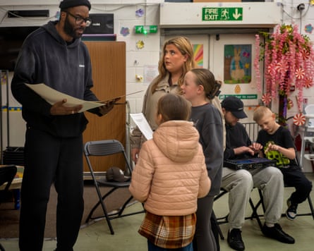 A tall young black man holding paper as he talks to a young woman and two small girls. Behind them are a young man and a boy using what looks like a synthesiser