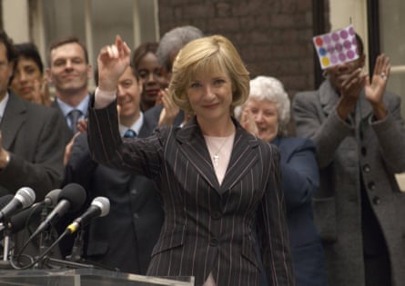 A woman with blond bobbed hair and pinstriped suit waving at camera with crowd of suits behind