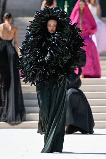 A fashion model walks down an outdoor runway wearing a dramatic black feathered couture garment