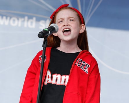 A young girl wears a red hoodie and red headband as she sings on stage.