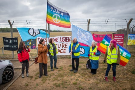 a group of people stand in front of the wire fencing holding flags including rainbows with ‘peace’ and the the CND symbols. There are banners on the fencing.