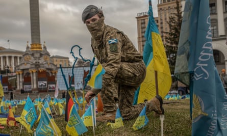 A soldier kneels on an area of grass where small Ukrainian flags have been planted