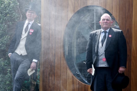Two men in morning suits stand amid the cooling mist emitted from a large fan