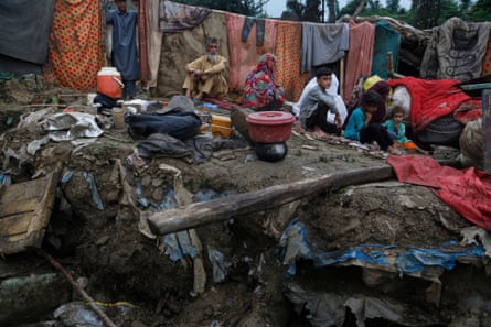 A family sits on muddy ground among scattered belongings. Behind them, a washing line is hung with sodden blankets and clothes