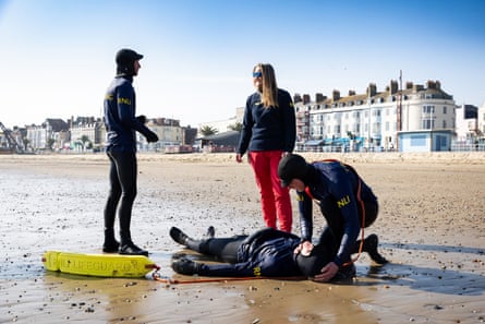 Alice on the beach with three male lifeguards, one of whom is lying on his back pretending to be someone who has been rescued