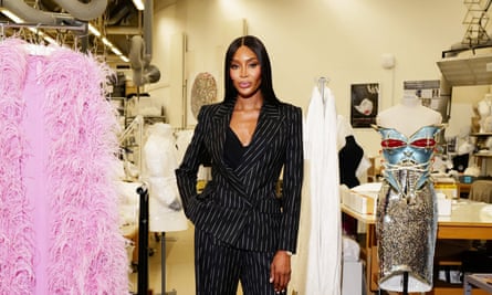 Naomi Campbell poses in a black suit surrounded by clothing.