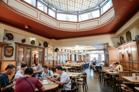 A room with vaulted skylight, wooden panelling and metal pans on its walls is filled with people sitting at wooden tables and chairs