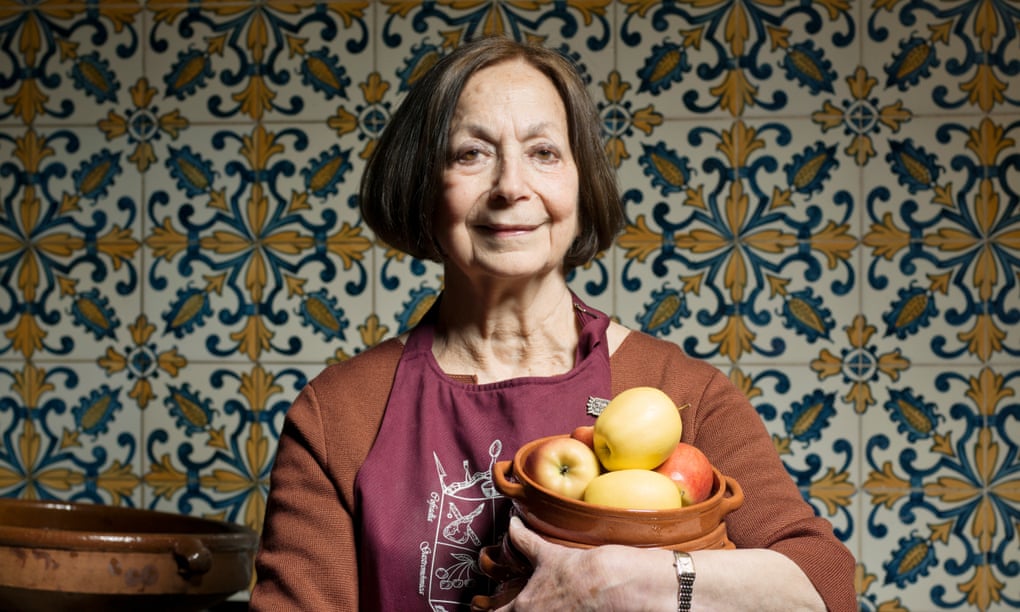 Claudia Roden at her home in an apron, a tiled kitchen wall behind, and holding a bowl of fruit