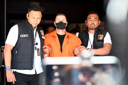 Steven Lyons in orange with a black mask is escorted by two police officers at the Bali police headquarters