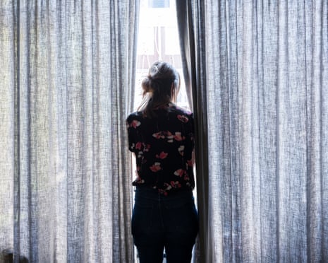 A woman in shadow parts curtains to look out window.