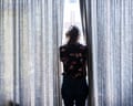 Photograph from behind of a woman looking out of a window, with long curtains