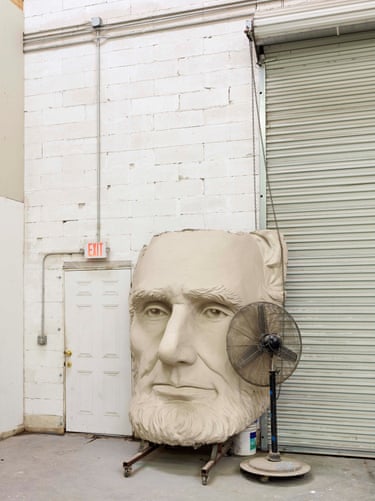 A giant sculpted presidential head sits in a storage yard beside an electric fan