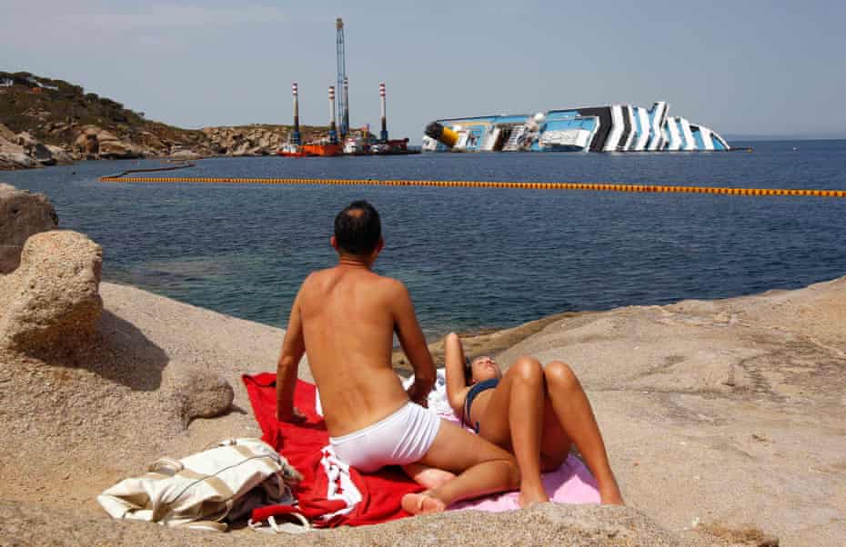 A couple sunbathe in front of the wreckage of the Costa Concordia near the harbour of Giglio Porto on 20 June 2012. Thirty-two people were killed when the cruise liner capsized after hitting rocks.