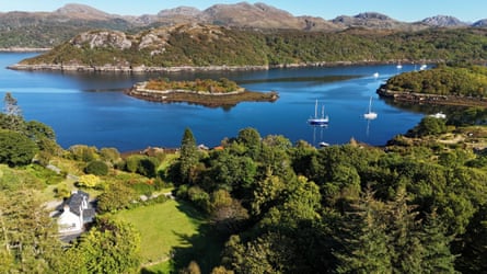 An aerial view of Arrowdale, a white cottage with a slate roof next to a loch with boats and islands in it and hills behind
