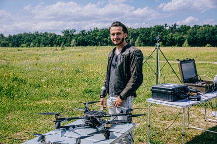 A man with a beard and wearing a leather jacket stands between two tables set up in a field with drone equipment on them